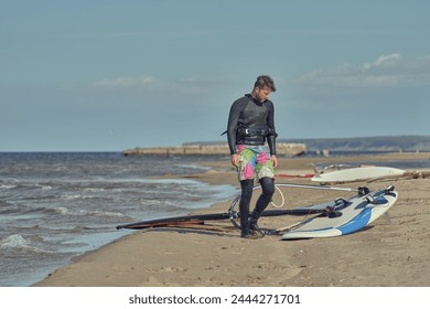 A mature man in a wetsuit, a windsurfer, stands on a sandy shore near a sailboard and rests. - Powered by Shutterstock