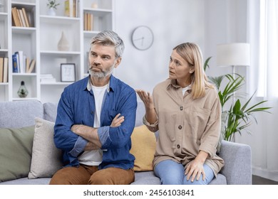 A mature man and woman having a serious conversation in a bright, modern living room. The man is upset, folding his arms, while the woman seems to be explaining something. - Powered by Shutterstock