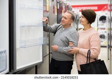 Mature Man And Woman Standing In Salesroom Of Appliance Shop And Choosing Refrigerator.