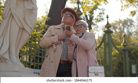 Mature man and woman tourists in stylish clothes standing near statue in park and reading information on cellphone. Aged couple walking in city park admiring sculptures