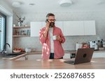 Mature man working from home on laptop and making phone call. Man standing by kitchen island, with cup of coffee. Concept of remote work from home. Hygge at work.