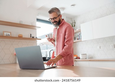 Mature man working from home on laptop, standing by kitchen island, with cup of coffee, holding smartphone. Concept of remote work from home. Hygge at work. - Powered by Shutterstock