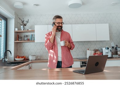 Mature man working from home on laptop and making phone call. Man standing by kitchen island, with cup of coffee. Concept of remote work from home. Hygge at work. - Powered by Shutterstock