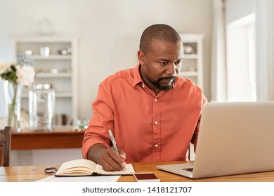 Mature man working on laptop while taking notes. Businessman working at home with computer while writing on agenda. African man managing home finance, reviewing bank account and using laptop.
