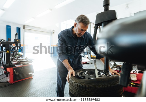 Mature man working in tire service\
workshop. Mechanic removing tire from wheel on\
machine.