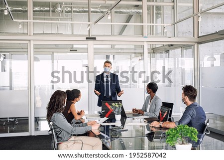 Mature manager in office discussing strategy with employees while wearing face mask during covid-19 pandemic. Group of multiethnic business people in meeting keeping social distancing in boardroom. 