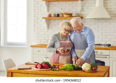 Mature Married Couple In Aprons Together Prepare Dinner At Home In A Bright Modern Kitchen. Loving Senior Man Helps His Wife Cut Vegetables For A Salad. Concept Of Seniors And Healthy Cooking.