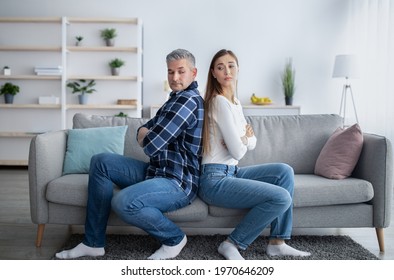 Mature married man and his wife sitting back to back on sofa, looking at each other, not knowing how to make peace after conflict, indoors. Middle-aged couple reconciling after family argument