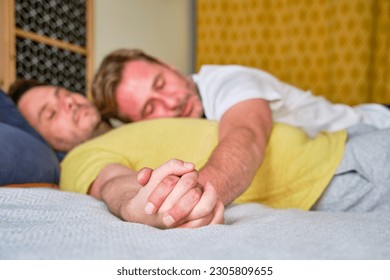 Mature married real gay couple lying in bed holding hands in foreground with unfocused faces in background - Powered by Shutterstock