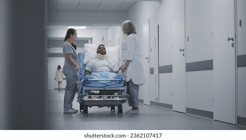 Mature medic and nurse walk down the clinic corridor with injured patient. Doctors transport African American man in neck brace on gurney to hospital ward. Medical staff at work in medical center. - Powered by Shutterstock