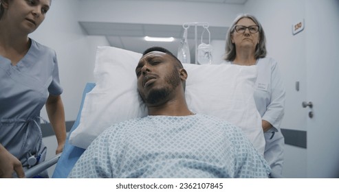 Mature medic with young nurse pushes middle aged patient on gurney to hospital ward. African American man lies on gurney, speaks with medics about his health. Medical staff at work in modern clinic. - Powered by Shutterstock