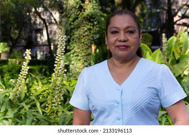 Mature Mexican lady looking at the camera smiling, standing in a park, with out of focus background of trees and green foliage. Latin woman looking at camera smiling. cheerful attitude.