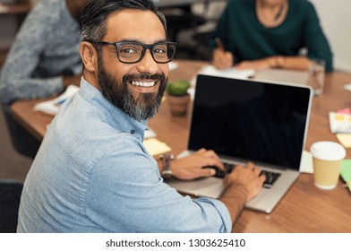 Mature middle eastern businessman using laptop at desk in busy office.Happy smiling casual man looking at camera while sitting at table and working on laptop computer. Indian leader using laptop.