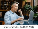 Mature middle-aged caucasian writer author writing novel book, businessman freelancer working remotely, e-learning using laptop in cafe restaurant