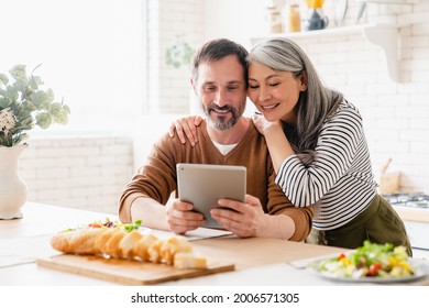 Mature middle-aged family couple wife and husband parents using digital tablet while having breakfast in the kitchen at home, reading news, searching web, reading e-book, e-learning remotely. - Powered by Shutterstock