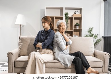 A mature mom and her young daughter, both Asian, sit on a sofa, engaged in a quarrel. Their argument escalates into a conflict, with clashes and friction over misunderstandings. - Powered by Shutterstock