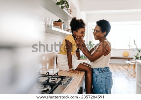 Mature mom stands in front of her daughter in the kitchen, smiling and holding her face lovingly. Happy black mom creating a special bond and an unbreakable relationship with her child.