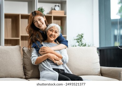 A mature mom and a young Asian woman, mother and daughter, sit together on a sofa in their living room, celebrating Mother's Day with love and bonding at home. - Powered by Shutterstock