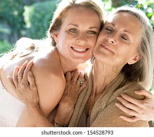 Mature Mother And Adult Daughter Spending Time Together In A Home Garden Hugging Tightly And Affectionately Cuddling During A Bright And Golden Summer Sunny Day Outdoors.