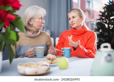 Mature mother and her adult daughter, who came to visit her for Christmas, drink tea and chat while sitting at a table in ..the room - Powered by Shutterstock