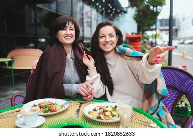 Mature mother and her young daughter sit together in cafe or restaurant. Cheerful woman pointing right forward. Look together in one direction. Having lunch outside at restaurant.
