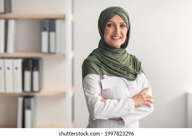 Mature Muslim Doctor Woman Wearing Hijab Smiling To Camera Standing Crossing Hands In Clinic Office. Portrait Of Professional Therapist Arab Lady Concept. Free Space For Text