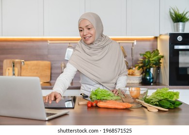 Mature muslim islamic woman in hijab cooking fresh vegetable salad using laptop for searching recipes online in the kitchen. Vegetarian and vegan food - Powered by Shutterstock