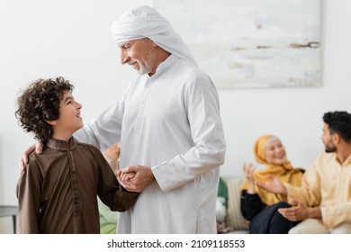 mature muslim man holding hand of arabian grandson near blurred interracial family at home