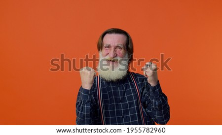 Mature old bearded gray-haired grandfather in shirt shouting, raising fists in gesture I did it, celebrating success, winning and goal achievement. Senior man on orange background. People lifestyle