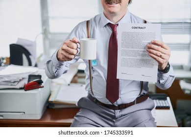 Mature old fashioned businessman holding lucrative contract and a cup of tea in hands with his office desk on background