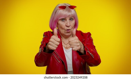 Mature old granny grandmother with pink hair in casual red jacket showing thumbs up and nodding in approval, successful good work. Senior woman rocker isolated on yellow background. People lifestyle
