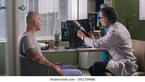 Mature oncologist explaining condition of incurable brain cancer to bald patient while sitting near computer during chemotherapy