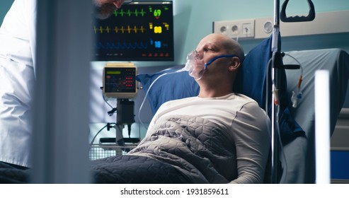 Mature Oncologist Putting On Respirator Mask And Pulse Oximeter On Bald Man And Turning On Oxygen Machine In Modern Ward Of Clinic