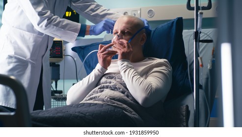 Mature Oncologist Putting On Respirator Mask And Pulse Oximeter On Bald Man And Turning On Oxygen Machine In Modern Ward Of Clinic