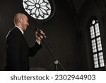 Mature pastor in black suit speaking prayer in microphone while standing in old baptist church