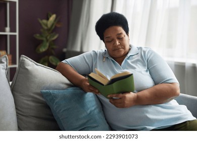 Mature plus size African American woman in casualwear sitting on couch with pillows and reading book while staying home on weekend - Powered by Shutterstock