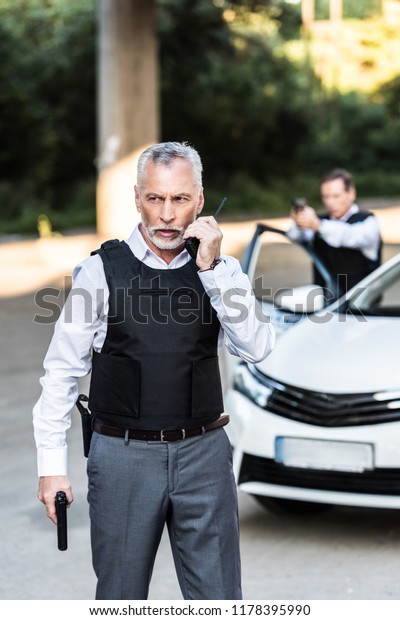 mature policeman with\
handgun talking on radio set while his colleague aiming by gun\
behind at street 