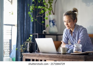 Mature Professional Asian Woman Working From Home Sitting In Couch Looking At Laptop Computer