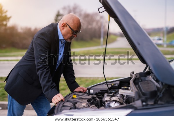 Mature professional elegant worried\
businessman in the suit is looking under the car hood trying to\
figure out the problem while leaning against the car.\
