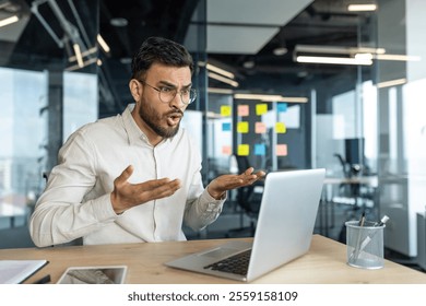 Mature professional man shows visible frustration using a laptop at his office desk. He appears in a collaborative office environment with glass walls and organized notes. - Powered by Shutterstock