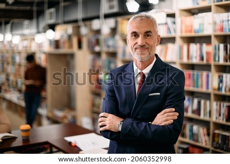 Mature professor standing with arms crossed in a library and looking at camera. 