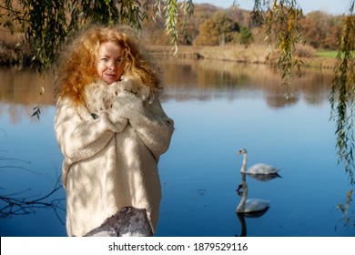 Mature Redheaded Woman In Her Best Years With Very Curly Hair Under A Weeping Willow Tree In Autumn At A Lake With Two Swimming Swans, Copy Space