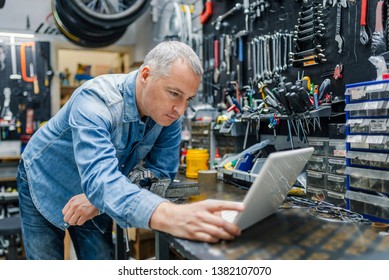 Mature Repairman with laptop. Male Carpenter Standing In His Workshop while looking at laptop. Mature Bike shop owner with laptop - Powered by Shutterstock