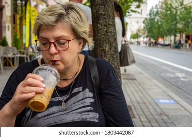 A Mature Sad Woman With Glasses Walks Through The Streets Of The Old City. Drinks A Cocktail At A Table In A Street Cafe