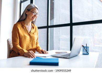 A mature satisfied asian gray-haired business woman wearing stylish casual clothes eyeglasses is using laptop for remote work, distance learning, online teaching