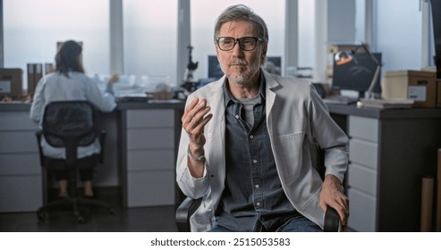 Mature scientist in lab coat speaks to the camera and gives interview for popular science film sitting in archaeological scientific laboratory. Female colleague works in the background. Static Shot. - Powered by Shutterstock - Get 15% off with code: PIKWIZARD15