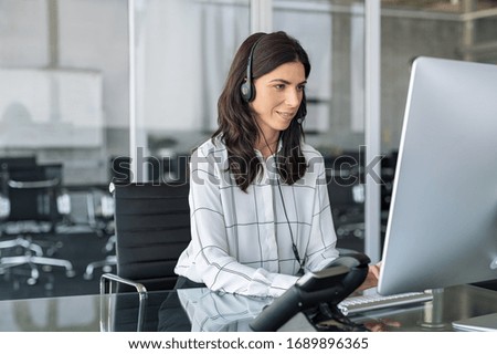 Mature secretary working in call center with headset. Confident telephone operator in modern office working on computer while taking calls. Smiling customer support operator at work with copy space.