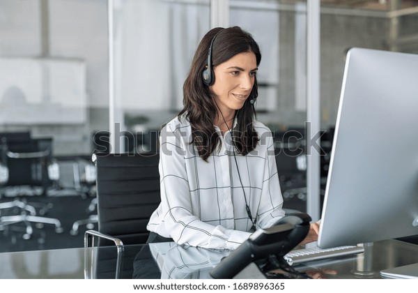 Mature secretary working in call center with\
headset. Confident telephone operator in modern office working on\
computer while taking calls. Smiling customer support operator at\
work with copy space.