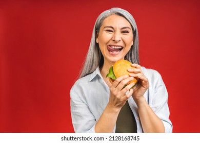 Mature Senior Asian Woman Eating Burger With Satisfaction. Grandmother Enjoys Tasty Hamburger Takeaway, Order Fastfood Delivery While Hungry, Standing Isolated Over Red Background.