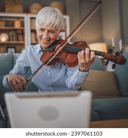 Mature senior caucasian woman learn to play violin practice at home online professor teacher teach or have internet private class on digital tablet while sit on sofa bed at home active senior hobby - Powered by Shutterstock